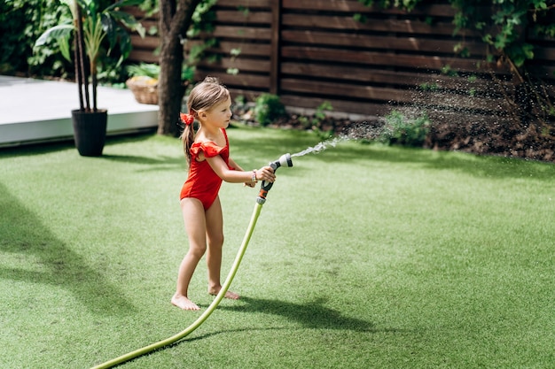 La petite fille mignonne dans un maillot de bain rouge arrose la pelouse La petite fille verse l'eau d'un tuyau d'arrosage