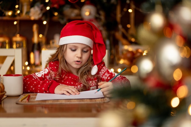 Une petite fille mignonne dans un pull rouge et un chapeau dans une cuisine sombre avec un arbre de Noël écrit une lettre au Père Noël et attend le nouvel an ou Noël