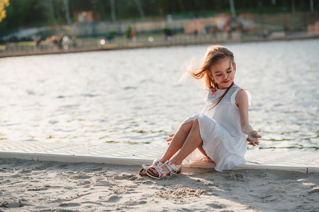 Une petite fille mignonne dans une robe blanche s'assied sur le sable sur l'heure d'été de couleurs chaudes de plage