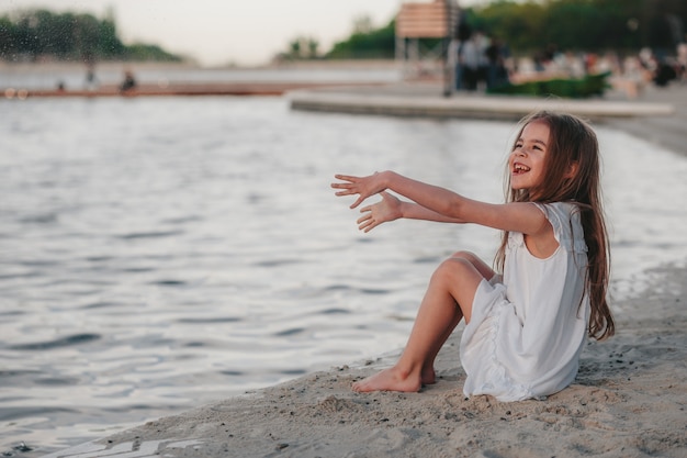 Une petite fille mignonne dans une robe blanche s'assied sur le sable sur l'heure d'été de couleurs chaudes de plage