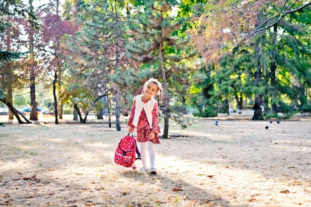 une petite fille mignonne dans une robe rouge une écolière se promène dans un parc en automne avec un sac à dos