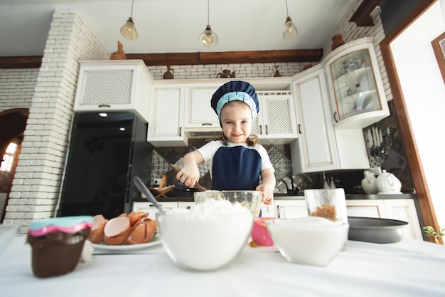 Une petite fille mignonne dans un tablier et un chapeau de chef remue la pâte avec une spatule en bois