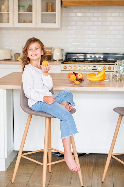 La petite fille mignonne dans des verres s'assied dans la cuisine près de la table avec des fruits et tient un abricot dans ses mains