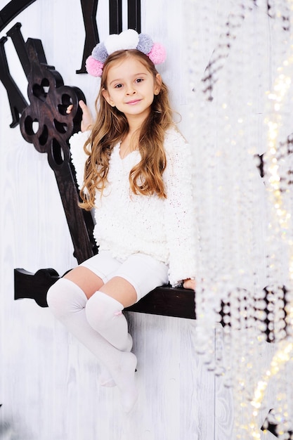 Une petite fille mignonne dans des vêtements légers et élégants est assise sur la main d'une énorme horloge à la veille du nouvel an ou à Noël.