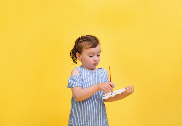 Une petite fille mignonne dessine avec une brosse avec un fouet dans ses mains sur un fond jaune avec un espace pour le texte.