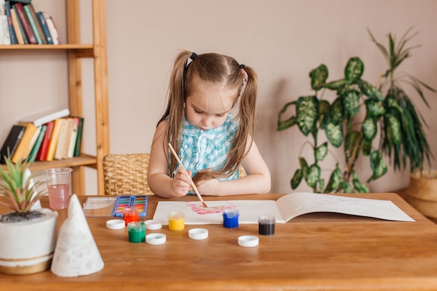 La petite fille mignonne dessine avec une brosse et peint à la table dans le salon