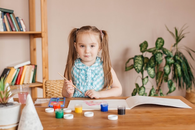 La petite fille mignonne dessine avec une brosse et peint à la table dans le salon