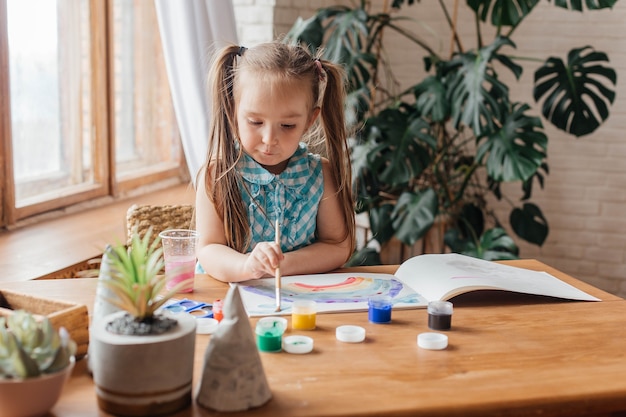 La petite fille mignonne dessine avec une brosse et peint à la table dans le salon