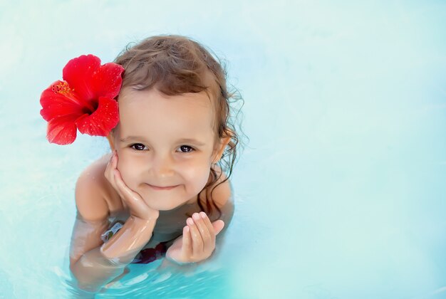 Petite fille mignonne enfant avec une fleur rouge derrière son oreille assis dans une eau bleue