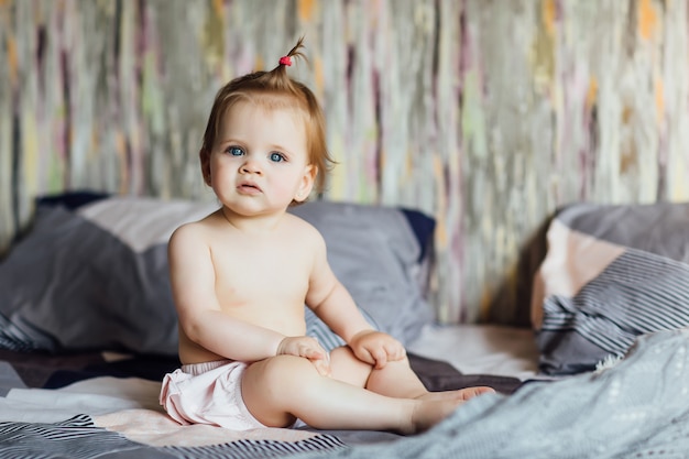 Petite fille mignonne est assise sur le lit avec une belle coiffure dans la chambre. Maison et mode de vie.