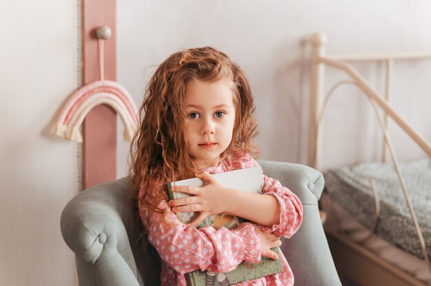 Une petite fille mignonne est assise avec un livre sur une chaise à l'intérieur des enfants