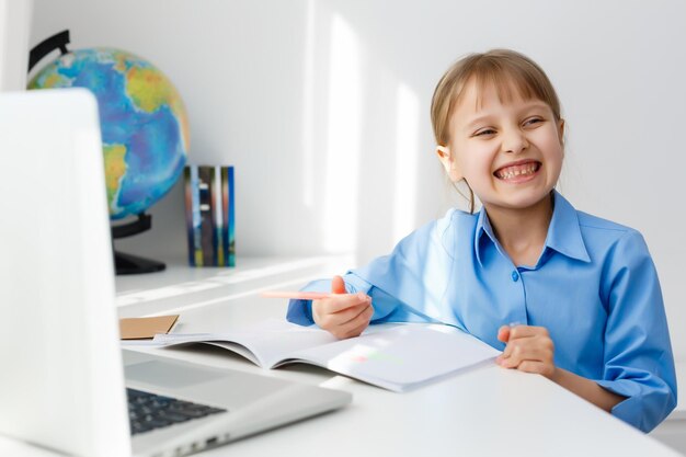 Une petite fille mignonne est assise à table avec son ordinateur portable et étudie en ligne