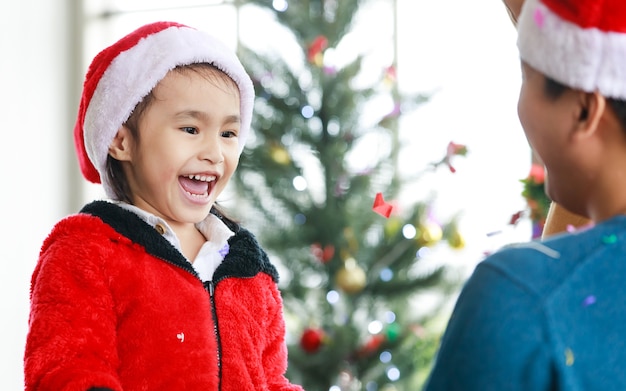 La petite fille mignonne de la famille asiatique heureuse porte un pull avec un chapeau de père Noël rouge et blanc debout en riant de façon excitante lorsque le père tire des confettis en papier brillant éclatant pour célébrer Noël.