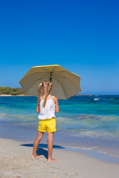 Petite fille mignonne avec grand parapluie jaune marchant sur la plage tropicale