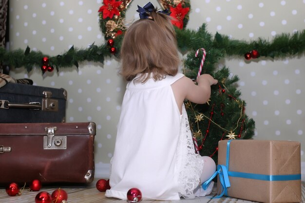 La petite fille mignonne habille l'arbre de Noël sur le sol dans la chambre