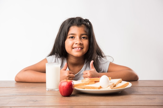 Petite fille mignonne indienne ou asiatique prenant son petit déjeuner. Table pleine de tranches de pain avec confiture, verre de lait, pomme fraîche et œuf dur
