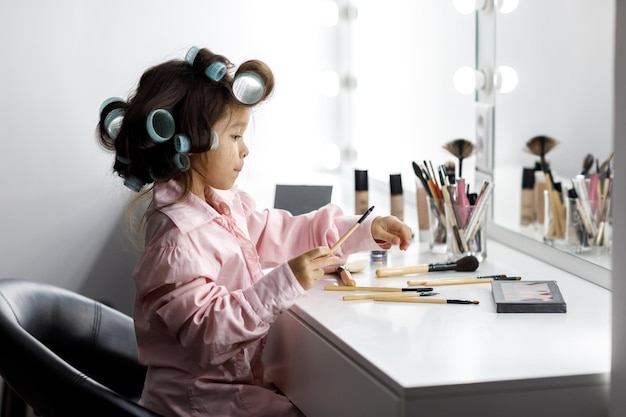 Petite fille mignonne jouant avec le cosmétique de sa mère devant le miroir à la maison