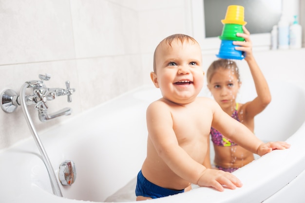 Petite fille mignonne jouant avec son petit frère dans la salle de bain avec de l'eau et différents jouets