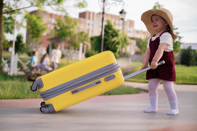 Une petite fille mignonne joyeuse dans une robe d'été bleue et un chapeau de paille tient une valise jaune