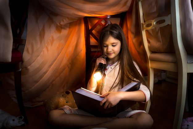 Petite fille mignonne lisant un livre dans une maison faite maison avec une lampe de poche