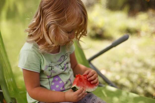 Photo petite fille mignonne mangeant la pastèque sur une chaise longue dans le jardin en été