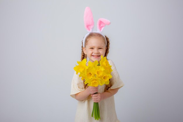 La petite fille mignonne avec des oreilles de lapin tient un bouquet des fleurs jaunes de jonquille sur un fond blanc