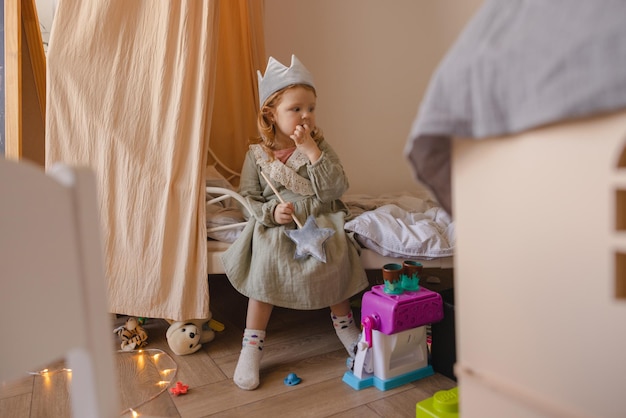 Petite fille mignonne à la peau claire en robe est assise sur le lit dans la chambre parmi les jouets Concept d'enfance