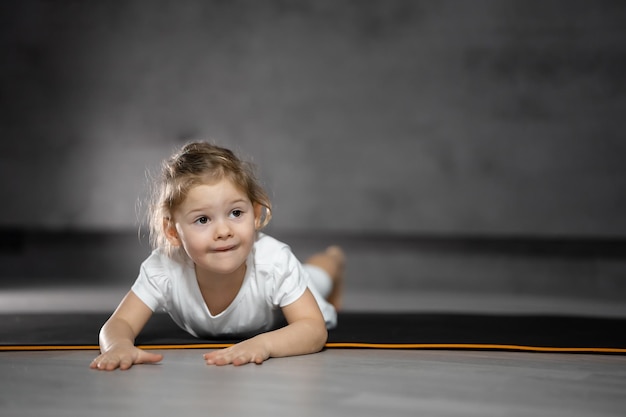 Petite fille mignonne pratiquant la pose de yoga sur fond gris dans une pièce sombre