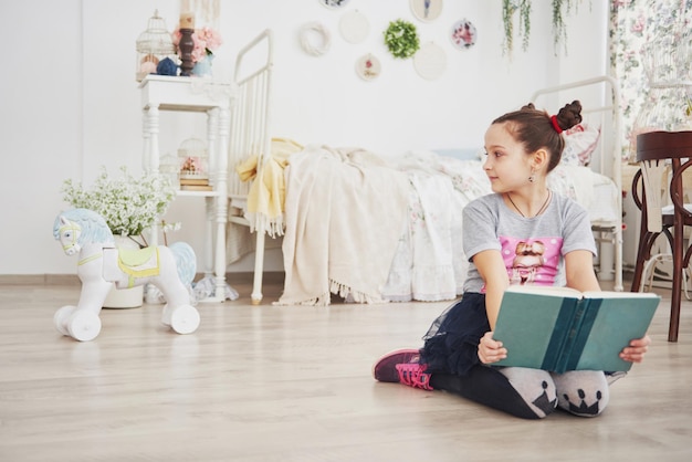 Une petite fille mignonne qui lit un livre dans la chambre, un enfant avec une couronne assis sur le lit près de la fenêtre.