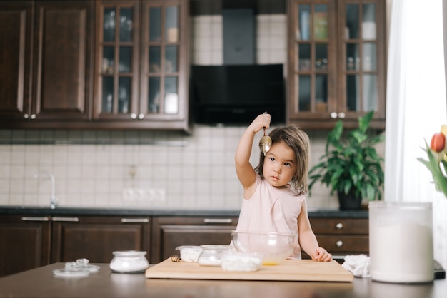 Une petite fille mignonne regarde la cuillère surélevée avec laquelle elle battait les œufs pour la pâte