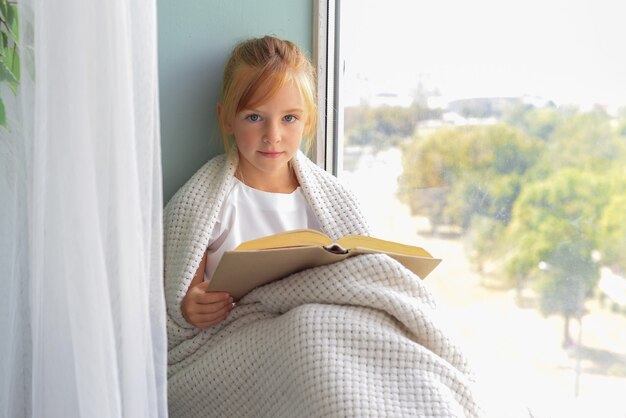 Une petite fille mignonne s'assied par la fenêtre et lit un livre dans la chambre à la maison Belle nature d'automne