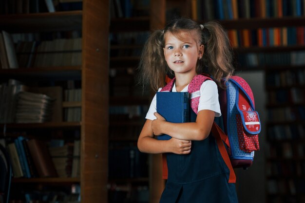 La petite fille mignonne avec le sac à dos se tient dans la bibliothèque pleine de livres