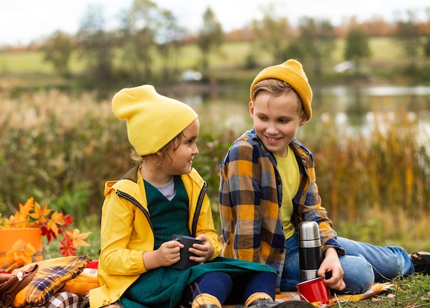 Petite fille mignonne souriante et garçon assis près de la voiture sur plaid et boire du cacao enfant se reposant avec sa famille dans la nature pique-nique saison d'automne