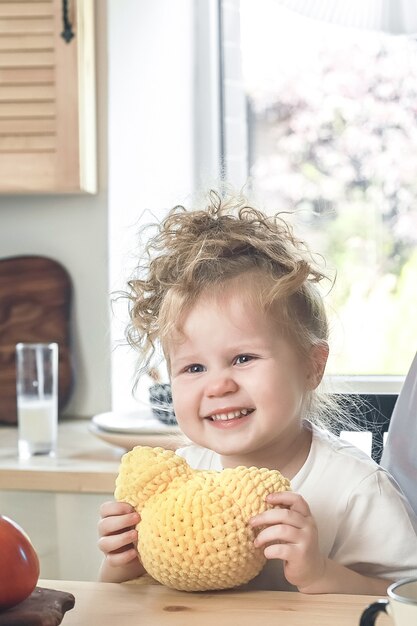 Petite fille mignonne souriante avec un jouet au crochet assis à table dans une cuisine lumineuse en attente de petit-déjeuner