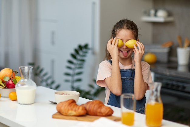 Petite fille mignonne souriante tenant des citrons près des yeux et montrant la langue sur fond flou de cuisine. Mode de vie sain et concept d'alimentation propre.