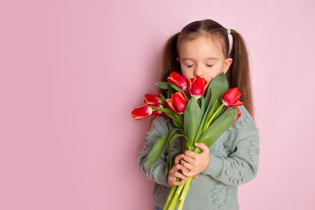 Une petite fille mignonne tenant un bouquet de tulipes et les reniflant avec les yeux fermés Fond rose. Joyeuse journée de la femme. Espace pour le texte. Émotions lumineuses. Photo de haute qualité