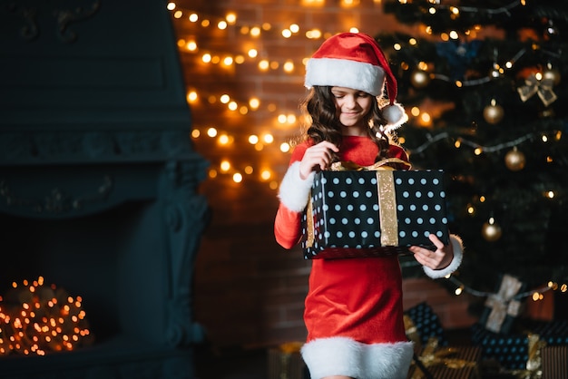 Petite fille mignonne tenant un cadeau près de l'arbre de Noël dans un costume de père Noël rouge