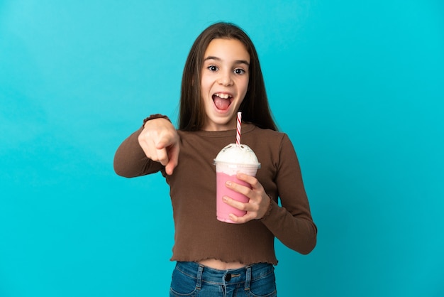 Petite fille avec milk-shake aux fraises isolé sur fond bleu surpris et pointant vers l'avant