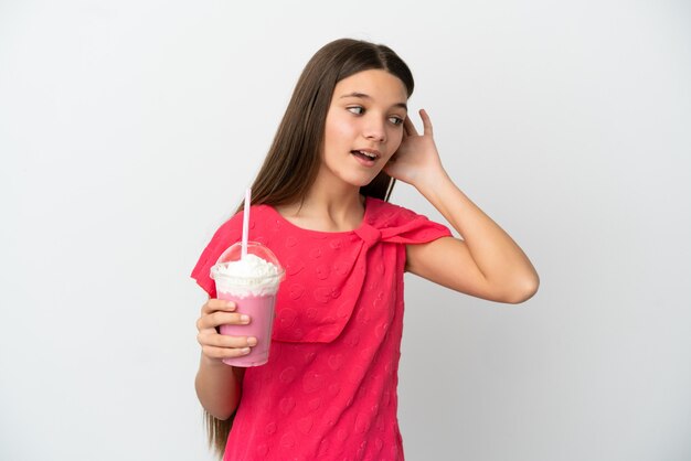 Petite fille avec milkshake aux fraises sur fond blanc isolé écoutant quelque chose en mettant la main sur l'oreille