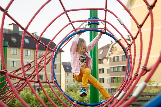 La petite fille monte des cordes au terrain de jeu sur fond de maisons en été