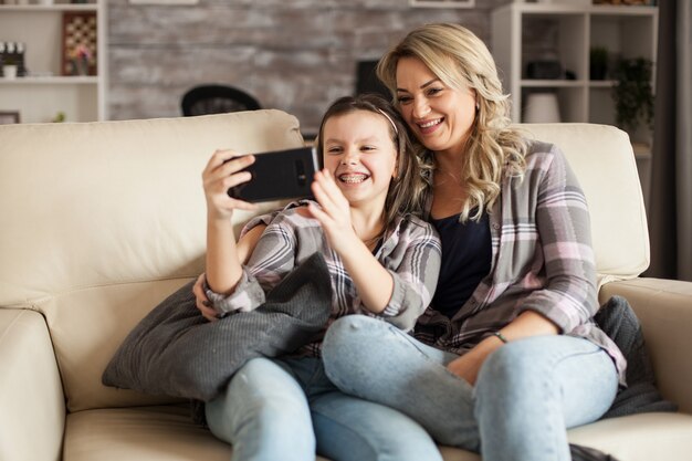 Petite fille montrant ses bretelles tout en prenant un selfie avec sa mère assise sur le canapé dans le salon.