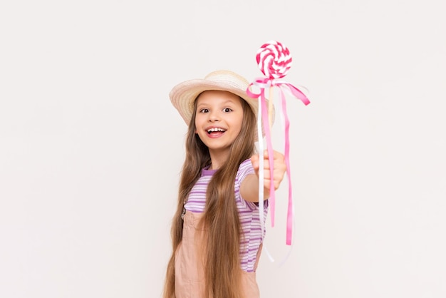 Une petite fille montre une sucette et sourit Un enfant dans une robe d'été et un chapeau d'été sur un fond blanc isolé