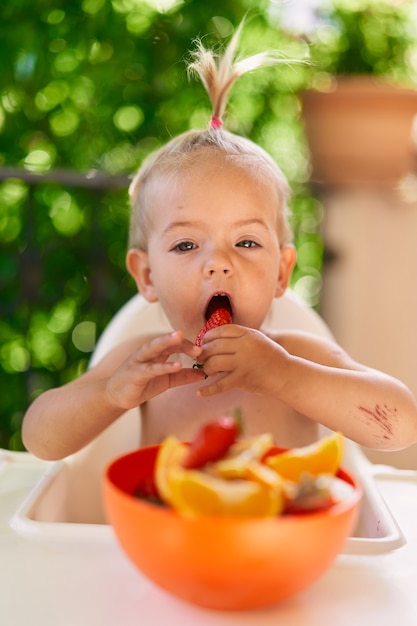 La petite fille mord la fraise tout en la tenant par ses mains