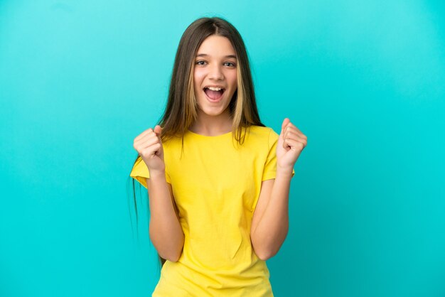 Petite fille sur un mur bleu isolé célébrant une victoire en position de vainqueur