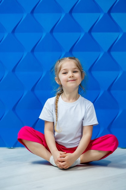 Petite fille avec des nattes dans un T-shirt blanc sur fond bleu. Elle est assise en position du lotus sur le sol et sourit et agite les mains.