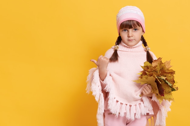 Petite fille avec des nattes habille un poncho rose chaud et une casquette avec des feuilles jaunes dans les mains