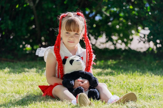 Petite fille avec des nattes rouges est assise sur l'herbe dans le parc avec un panda ours en peluche. photo de haute qualité