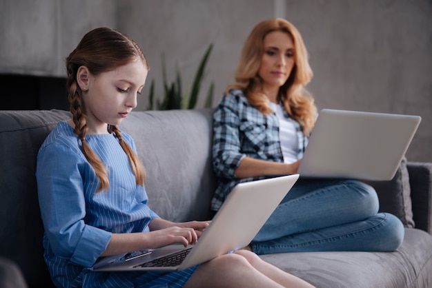 Petite fille obsédée mignonne assise à la maison et surfant sur Internet pendant que la mère travaille