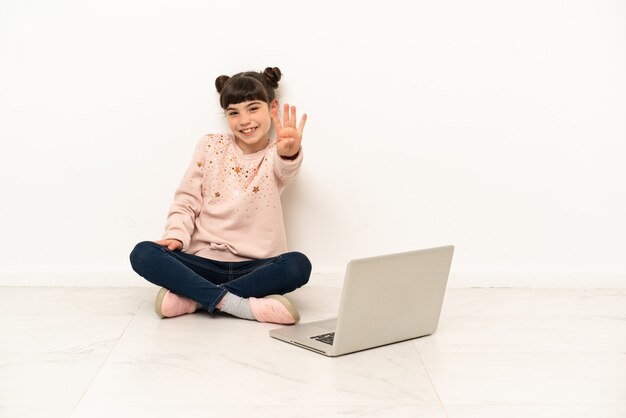 Petite fille avec un ordinateur portable assis sur le sol heureux et comptant quatre avec les doigts