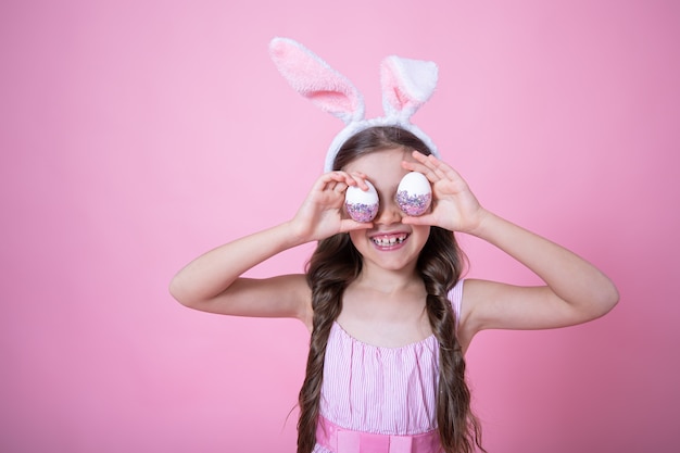 Petite fille avec des oreilles de lapin de Pâques posant tenant des oeufs de Pâques festifs dans ses mains sur fond rose studio close up